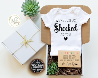 May include: A white onesie with the text "We're just as shocked as you!" and a black heart. A small card with the text "Could be pink, could be blue. All we know is that We Are Due!" is in front of the onesie. The onesie and card are in a brown box with shredded paper.