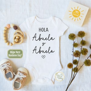 May include: White baby onesie with black text that reads "Hola Abuela y Abuelo" with a small heart symbol below the text.