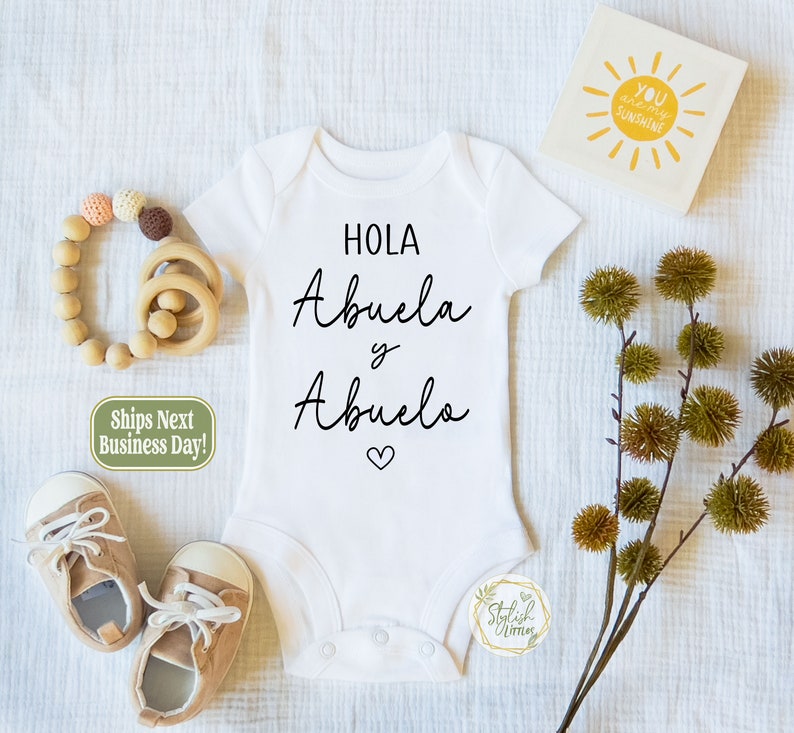 May include: White baby onesie with black text that reads "Hola Abuela y Abuelo" with a small heart symbol below the text.