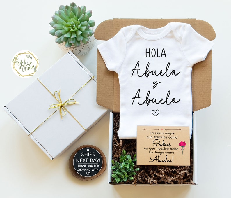 May include: A white baby onesie with the text "HOLA Abuela y Abuelo" in black. The onesie is in a gift box with a small card that reads "Lo unico mejor que tenerlos como Padres es que nuestro bebe los tenga como Abuelos!"