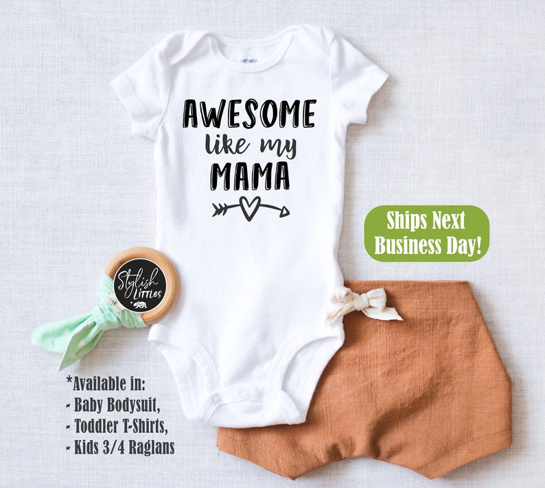 Op de afbeelding: Witte baby-romper met zwarte tekst die "AWESOME like my MAMA" luidt, met een pijl die naar een hart wijst. De romper is op een witte achtergrond.