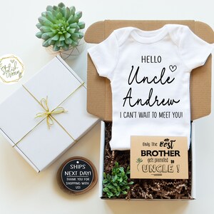 May include: A white onesie with the text "Hello Uncle Andrew I can't wait to meet you!"  A brown card with the text "Only the best brother gets promoted to uncle!"  A white gift box with a gold ribbon and a small wooden sign that says "Ships next day! Thank you for shopping with us!"