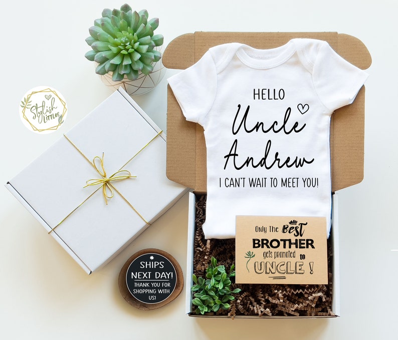 May include: A white onesie with the text "Hello Uncle Andrew I can't wait to meet you!"  A brown card with the text "Only the best brother gets promoted to uncle!"  A white gift box with a gold ribbon and a small wooden sign that says "Ships next day! Thank you for shopping with us!"