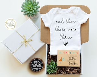 May include: A white onesie with the text "and then there were three" and a small heart with two footprints. The onesie is in a brown cardboard box with a white gift box, a card with a stork and the text "I've loved you as a Hubby & I'll adore you as a Daddy..." and two small potted succulents.