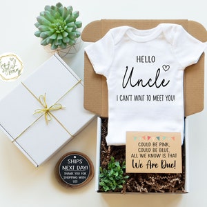 May include: A white onesie with the text "Hello Uncle I can't wait to meet you!"  A brown card with the text "Could be pink, could be blue, all we know is that We Are Due!"  A white gift box with a gold ribbon and a small wooden sign that says "Ships Next Day!"