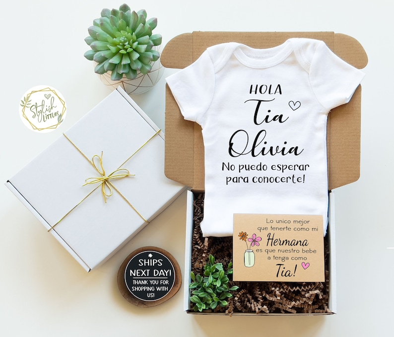 May include: A white onesie with the text "HOLA Tia Olivia No puedo esperar para conocerte!" in black. A small brown card with the text "Lo unico mejor que tenerte como mi Hermana es que nuestro bebe a tenga como Tia!" in black and pink. The onesie and card are in a brown box with a small green plant.