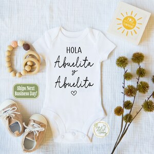 May include: White baby onesie with black text that reads "Hola Abuelita y Abuelito" with a small heart symbol. The onesie is surrounded by baby accessories, including a wooden teether, a pair of brown baby shoes, and a small green and white tag.