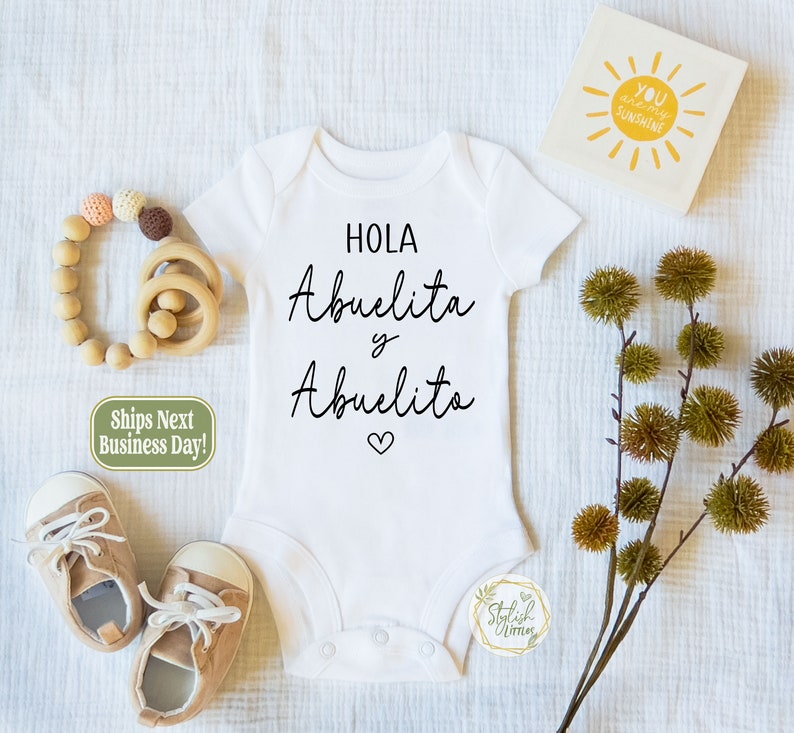 May include: White baby onesie with black text that reads "Hola Abuelita y Abuelito" with a small heart symbol. The onesie is surrounded by baby accessories, including a wooden teether, a pair of brown baby shoes, and a small green and white tag.
