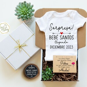 May include: A white onesie with the text "Sorpresa! Bebe Santos llegando Diciembre 2023" in Spanish. The onesie is in a brown box with a small card that reads "Lo unico mejor que tenerlos como Padres es que nuestro bebe los tenga como Abuelos!" in Spanish.