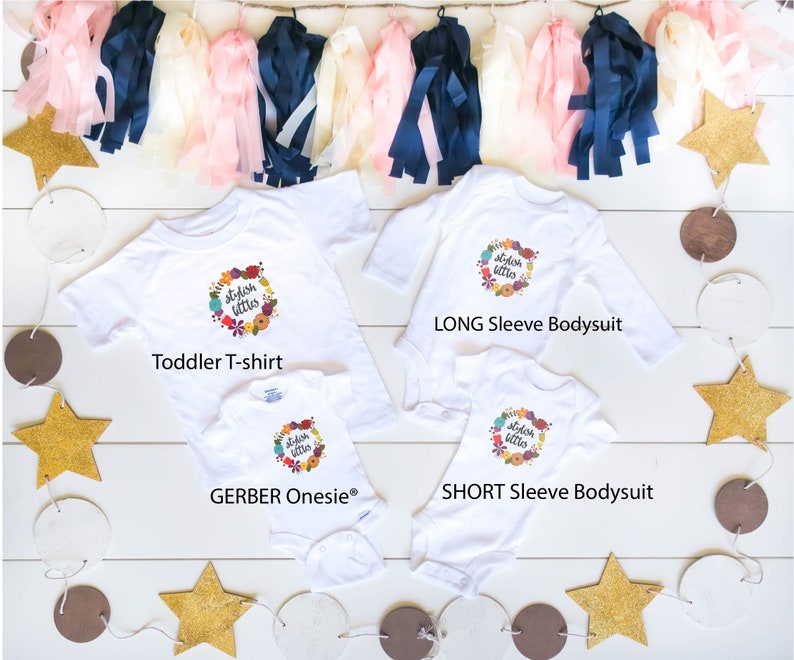 Może przedstawiać: Trzy białe koszulki z napisem "stylish littles" w wianku z kwiat&oacute;w. Koszulki to koszulka dla małych dzieci, body z długim rękawem i body z kr&oacute;tkim rękawem.