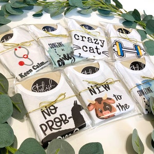 Op de afbeelding: Een verzameling van tien witte baby-rompers met verschillende ontwerpen en spreuken, waaronder "Someone Special", "Keen Calm", "Stylish", "No Prob", "Crazy Cat", "New to the", "Someone Special" en "Keep Calm". De rompers zijn verpakt in doorzichtige plastic zakken met een gouden lint.