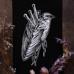 May include: A black and white illustration of a bird pierced by nails. The bird is depicted with detailed wing feathers and a beak, with blood droplets. The artwork is on a black background.