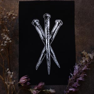 Puede incluir: Parche rectangular negro con una ilustración blanca de tres clavos dispuestos en forma triangular. Los clavos tienen detalles texturizados. El parche está sobre un fondo oscuro, con flores secas en primer plano.