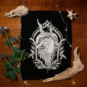 Può includere: Illustrazione in bianco e nero di una testa di capra con grandi corna, incorniciata da un bordo ornato. L'opera d'arte è su sfondo nero, con un bordo strutturato. Elementi decorativi: salvia, cardo e osso.