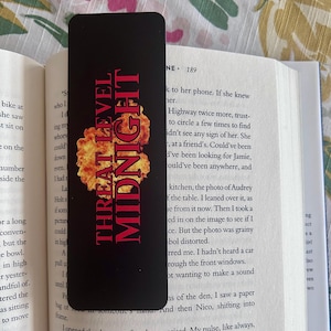 May include: Black bookmark with rounded corners featuring the text "THREAT LEVEL MIDNIGHT" in red and orange, with a fiery graphic. The bookmark is placed in an open book with white pages.