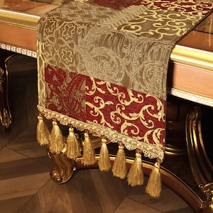 May include: A decorative table runner with a rich, ornate design. The runner features a combination of burgundy and gold colors with intricate patterns. Gold tassels hang from the bottom edge, adding a touch of elegance. The runner is likely made of a luxurious fabric.