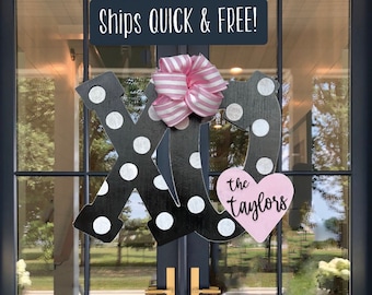 May include: Black and white wooden "XO" sign with white polka dots and a pink bow. A pink heart with the name "the Taylors" is attached to the "O". The sign is hanging on a dark blue door with gold handles.
