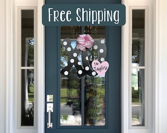 May include: A black and white polka dot door wreath with a pink bow and a pink heart with the text "the Taylors" hanging on a blue front door with a sign above that reads "Free Shipping".