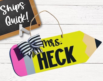 May include: A yellow pencil-shaped wooden sign with a pink and gray eraser. The sign says "mrs. HECK" in black letters with a black and white bow on top.