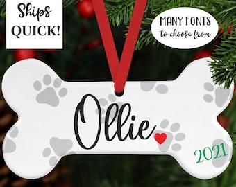 May include: White ceramic bone-shaped Christmas ornament with gray paw prints and the name "Ollie" written in black cursive. A red heart is next to the name and the year "2021" is written in green on the bottom right corner. The ornament is hanging from a red ribbon.