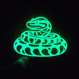 May include: A neon green snake-shaped object against a black backdrop. The snake is coiled, with detailed scales and a triangular head. The object emits a bright, neon-like light, creating a striking contrast.
