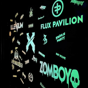 K&ouml;nnte beinhalten: Eine schwarze Wand mit neongr&uuml;nen und wei&szlig;en Logos und Texten f&uuml;r verschiedene K&uuml;nstler und Veranstaltungen. Der Text "FLUX PAVILION" ist prominent dargestellt, zusammen mit "ZOMBOY" und einer Totenkopf-Grafik. Weitere Logos sind "ILLENIUM" und "SEVEN LIONS".