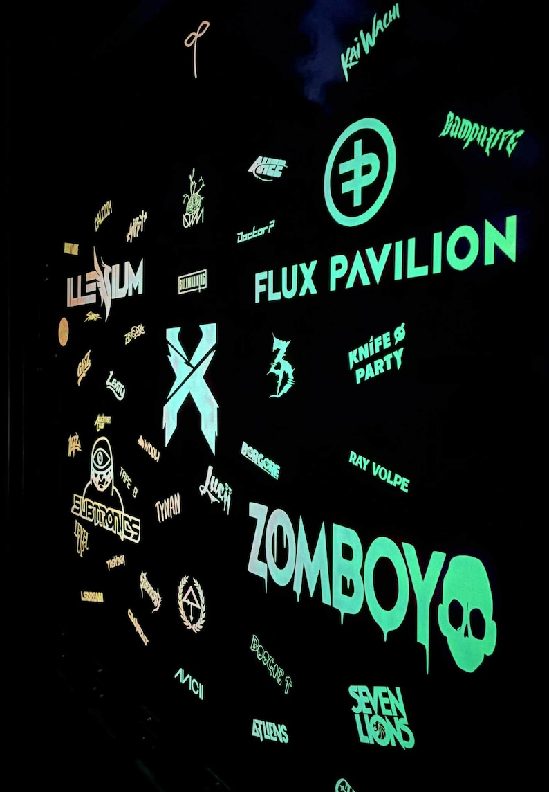 K&ouml;nnte beinhalten: Eine schwarze Wand mit neongr&uuml;nen und wei&szlig;en Logos und Texten f&uuml;r verschiedene K&uuml;nstler und Veranstaltungen. Der Text "FLUX PAVILION" ist prominent dargestellt, zusammen mit "ZOMBOY" und einer Totenkopf-Grafik. Weitere Logos sind "ILLENIUM" und "SEVEN LIONS".