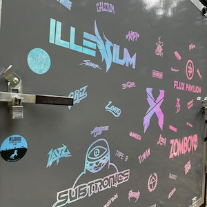K&ouml;nnte beinhalten: Eine graue Anh&auml;ngert&uuml;r, die mit holografischen Aufklebern bedeckt ist. Die Aufkleber zeigen verschiedene Logos und Texte, darunter "ILLENIUM", "SUBTRONICS" und "ZOMBOY". Andere Aufkleber zeigen Namen und Symbole.