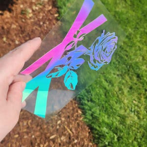 May include: A clear, iridescent sticker with a rose and the letter "X" in pink, purple, and teal. The floral design is detailed, with visible petals and leaves. The sticker is held against a backdrop of green grass and brown mulch.