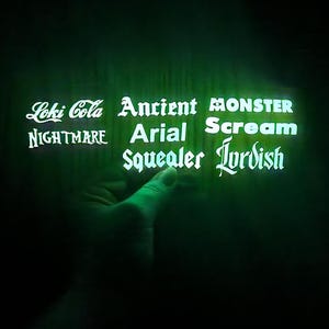 Może przedstawiać: Ciemnozielona, świecąca prostokątna powierzchnia z białym tekstem Loki Cola, Ancient, MONSTER, NIGHTMARE, Arial, Scream, Squealer oraz Iordish ułożonym w siatkę na czarnym tle.