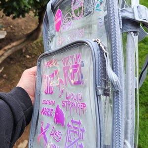 K&ouml;nnte beinhalten: Transparenter Rucksack mit grauem Rand und mehreren Rei&szlig;verschlussf&auml;chern. Die Vordertasche zeigt holografische Aufkleber mit Text und Grafiken. Der Rucksack hat verstellbare Tr&auml;ger und einen oberen Griff.