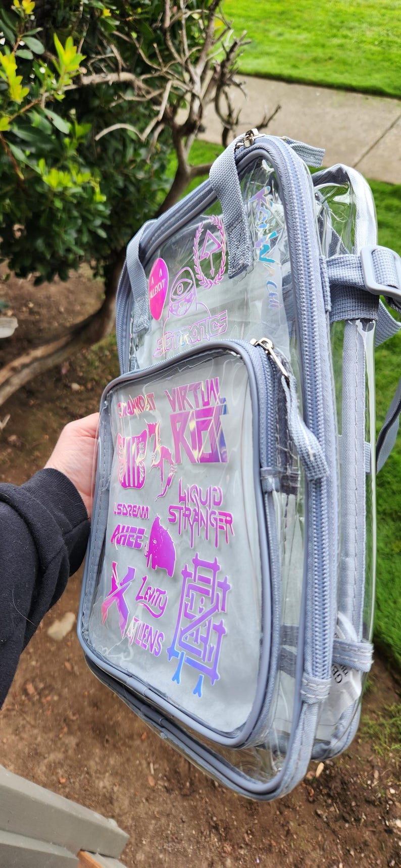 K&ouml;nnte beinhalten: Transparenter Rucksack mit grauem Rand und mehreren Rei&szlig;verschlussf&auml;chern. Die Vordertasche zeigt holografische Aufkleber mit Text und Grafiken. Der Rucksack hat verstellbare Tr&auml;ger und einen oberen Griff.