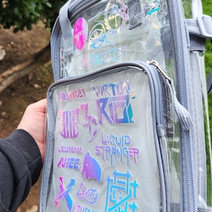 K&ouml;nnte beinhalten: Transparenter Rucksack mit grauem Besatz und mehreren Rei&szlig;verschlussf&auml;chern. Die Vordertasche hat holografische Aufkleber mit verschiedenen Texten und Designs. Der Rucksack hat graue Tr&auml;ger und Griffe.