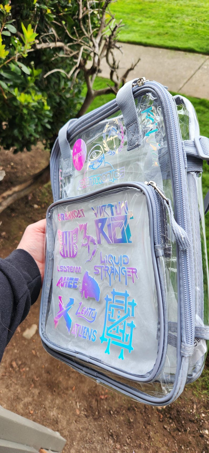 K&ouml;nnte beinhalten: Transparenter Rucksack mit grauem Besatz und mehreren Rei&szlig;verschlussf&auml;chern. Die Vordertasche hat holografische Aufkleber mit verschiedenen Texten und Designs. Der Rucksack hat graue Tr&auml;ger und Griffe.