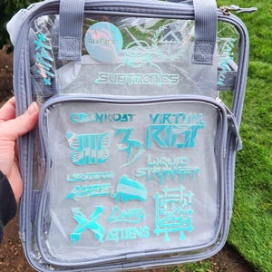 K&ouml;nnte beinhalten: Transparente Tragetasche mit grauem Besatz und Griffen. Die Tasche zeigt verschiedene t&uuml;rkisfarbene Logos und Texte, darunter "SUBTRONICS" und "CRANKDAT". Die Tasche wird vor einem gr&uuml;nen Rasen und einem Busch gehalten.