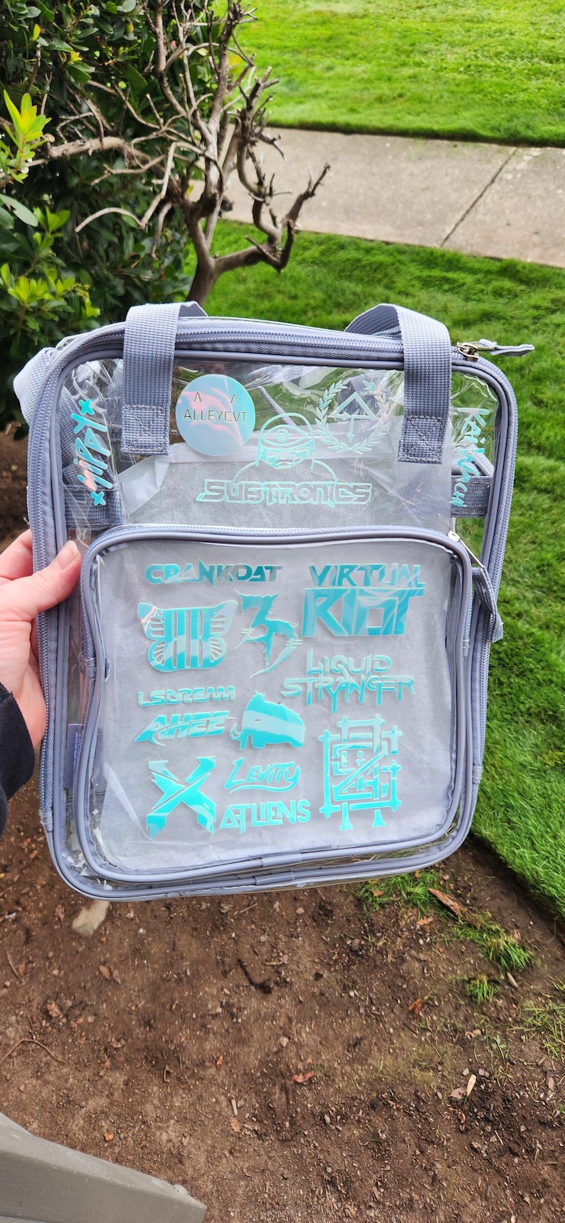 K&ouml;nnte beinhalten: Transparente Tragetasche mit grauem Besatz und Griffen. Die Tasche zeigt verschiedene t&uuml;rkisfarbene Logos und Texte, darunter "SUBTRONICS" und "CRANKDAT". Die Tasche wird vor einem gr&uuml;nen Rasen und einem Busch gehalten.