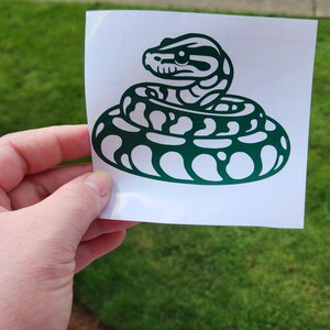 May include: A green snake sticker on a white background. The sticker features a coiled snake design with detailed scales and a triangular head. The sticker is held in a hand against a backdrop of green grass.