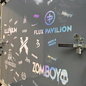 K&ouml;nnte beinhalten: Eine graue Anh&auml;ngert&uuml;r ist mit holografischen Aufklebern und Logos bedeckt. Die W&ouml;rter "FLUX PAVILION" und "ZOMBOY" sind deutlich sichtbar. Weitere Logos sind "ILLENIUM", "Knife Party" und "Seven Lions". Die T&uuml;r hat einen Metallgriff und einen Riegel.