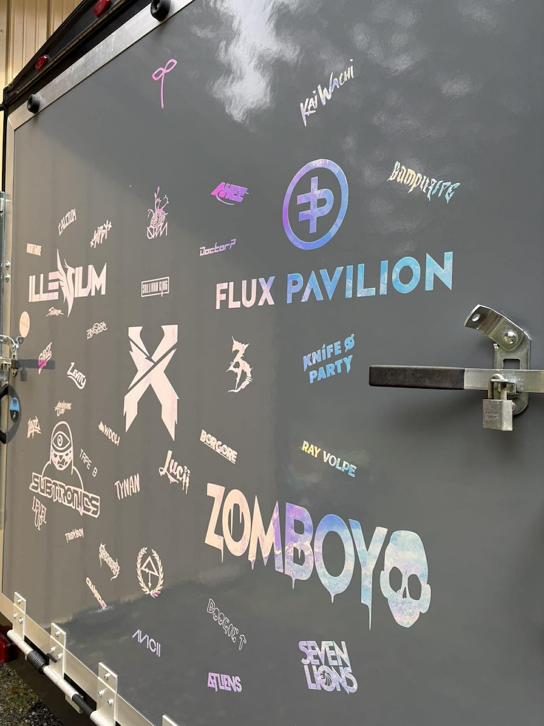 K&ouml;nnte beinhalten: Eine graue Anh&auml;ngert&uuml;r ist mit holografischen Aufklebern und Logos bedeckt. Die W&ouml;rter "FLUX PAVILION" und "ZOMBOY" sind deutlich sichtbar. Weitere Logos sind "ILLENIUM", "Knife Party" und "Seven Lions". Die T&uuml;r hat einen Metallgriff und einen Riegel.