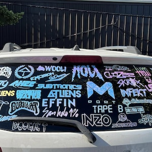K&ouml;nnte beinhalten: Heckscheibe eines wei&szlig;en Autos mit holografischen Aufklebern. Die Aufkleber zeigen Logos und Texte, darunter "WOOLI", "SUBTRONICS" und "INFEKT". Der Text ist in verschiedenen Schriftarten und Farben gehalten.