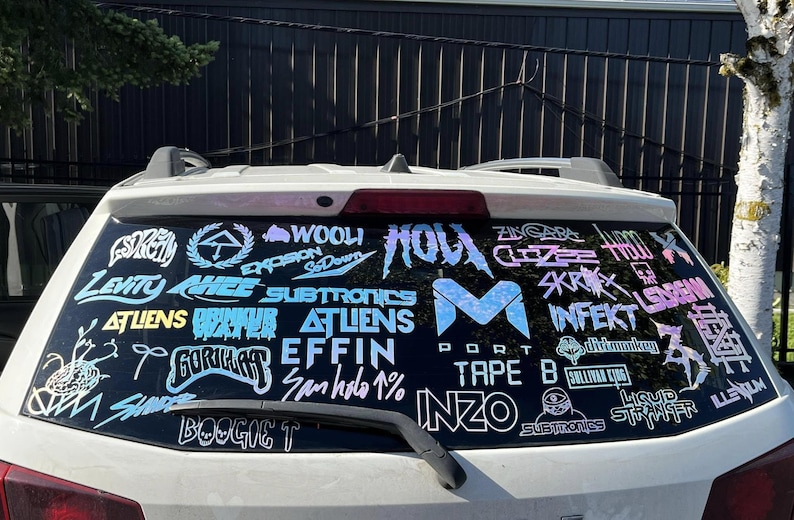 K&ouml;nnte beinhalten: Heckscheibe eines wei&szlig;en Autos mit holografischen Aufklebern. Die Aufkleber zeigen Logos und Texte, darunter "WOOLI", "SUBTRONICS" und "INFEKT". Der Text ist in verschiedenen Schriftarten und Farben gehalten.