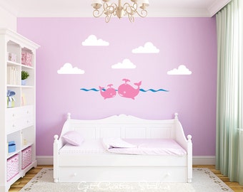 May include: A wall decal set featuring two pink whales swimming in blue waves, accompanied by five white clouds. The design is applied to a light purple wall above a white daybed, creating a cohesive decorative arrangement for a room interior.