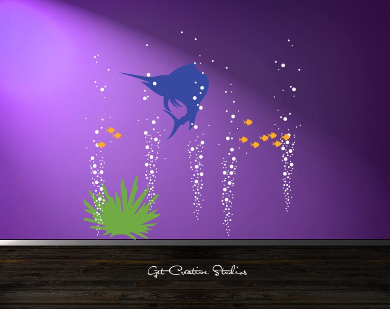 May include: A wall decal set featuring a blue sailfish silhouette, a green sea plant, small orange fish, and white bubble streams. The design is displayed on a purple wall above a dark wooden floor with the text Get Creative Studios at the bottom.