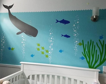 May include: A wall decal set featuring a large gray whale, various blue and green fish, white bubble streams, and green seaweed on a light blue background. The design includes a scalloped white border at the top edge of the wall mural.