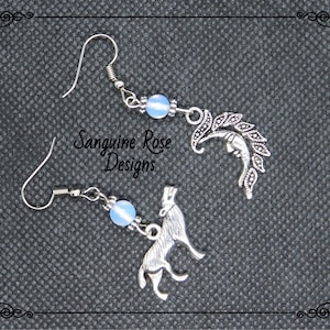 May include: A pair of silver earrings with blue glass beads. One earring features a crescent moon with leaves, the other a wolf howling at the moon.