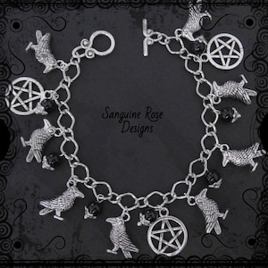 Op de afbeelding: Een zilveren armband met bedels met zwarte kralen en bedels van raven en pentagrammen. De armband heeft een kreeft sluiting en een schuifsluiting. De tekst "Sanguine Rose Designs" is zichtbaar op de armband.