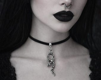 May include: A black velvet choker necklace featuring a silver-toned snake pendant with textured scales. The pendant hangs vertically from a simple silver bail attached to the thin black band, resting against the neck.