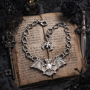 Op de afbeelding: Een zilverkleurige armband met vleermuisthema, met een grote vleermuisbedel en kleinere vleermuisbedels. De armband wordt getoond op een open antiek boek met gotische accessoires.
