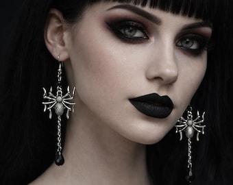 May include: A pair of silver-toned spider earrings featuring a detailed arachnid design with eight legs. Each earring hangs from a hook and includes a short silver chain dangling from the spider's body, ending in a small, smooth black teardrop bead.