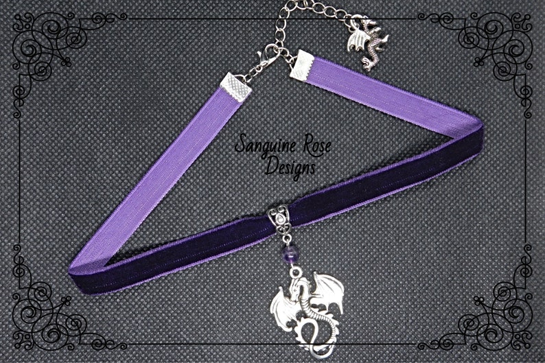 May include: A purple velvet choker necklace with a silver dragon pendant. The pendant has a purple gemstone and is attached to a silver chain. The choker is adjustable and has a silver clasp.